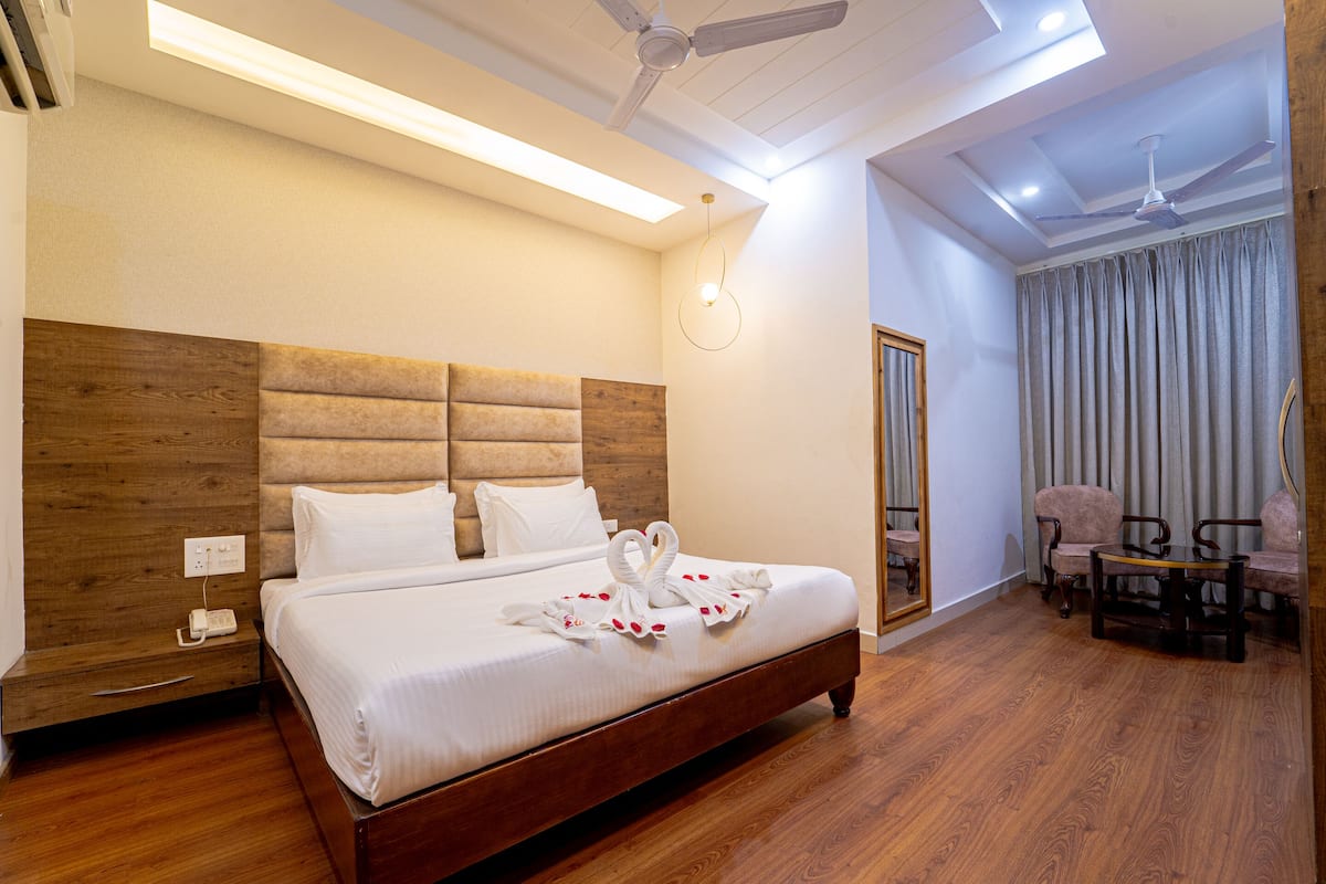 Une chambre d’hôtel avec une tête de lit en bois, un lit avec du linge de maison blanc, un coin repas avec une table et des chaises, et un ventilateur de plafond.