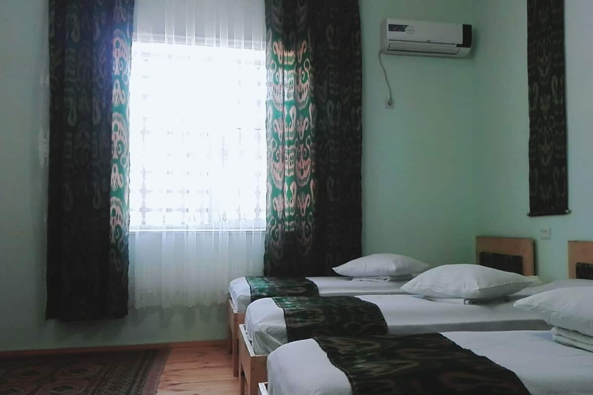 A room with three single beds, a window with patterned curtains, an air conditioning unit, and a wooden floor.