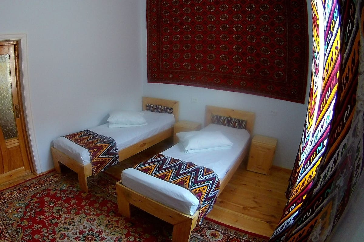 A room with two single beds, a wooden door, a patterned rug, and a red wall tapestry.