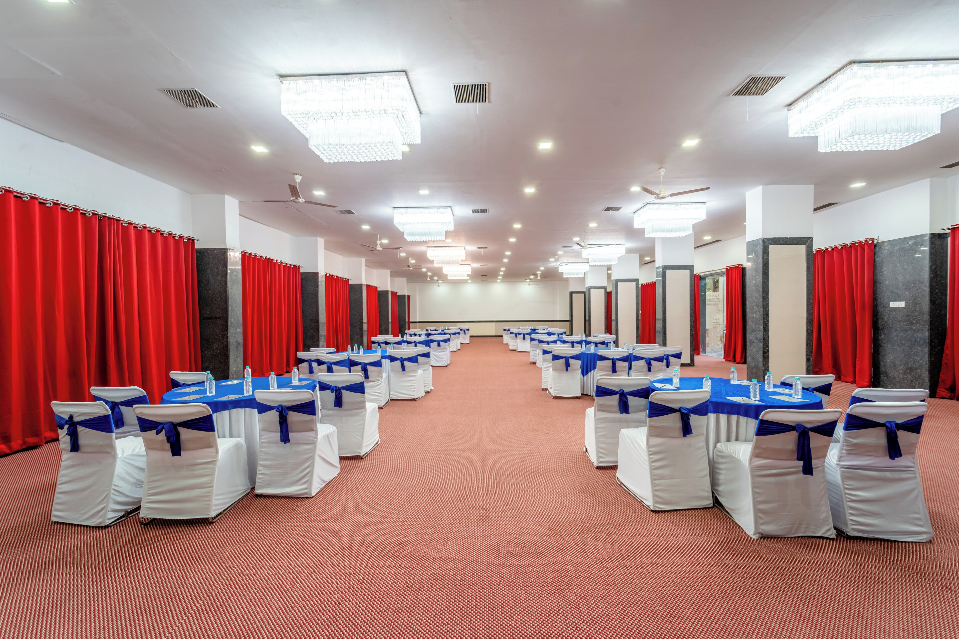 Un salón de conferencias con filas de sillas blancas con cintas azules, cortinas rojas y mesas con manteles azules.