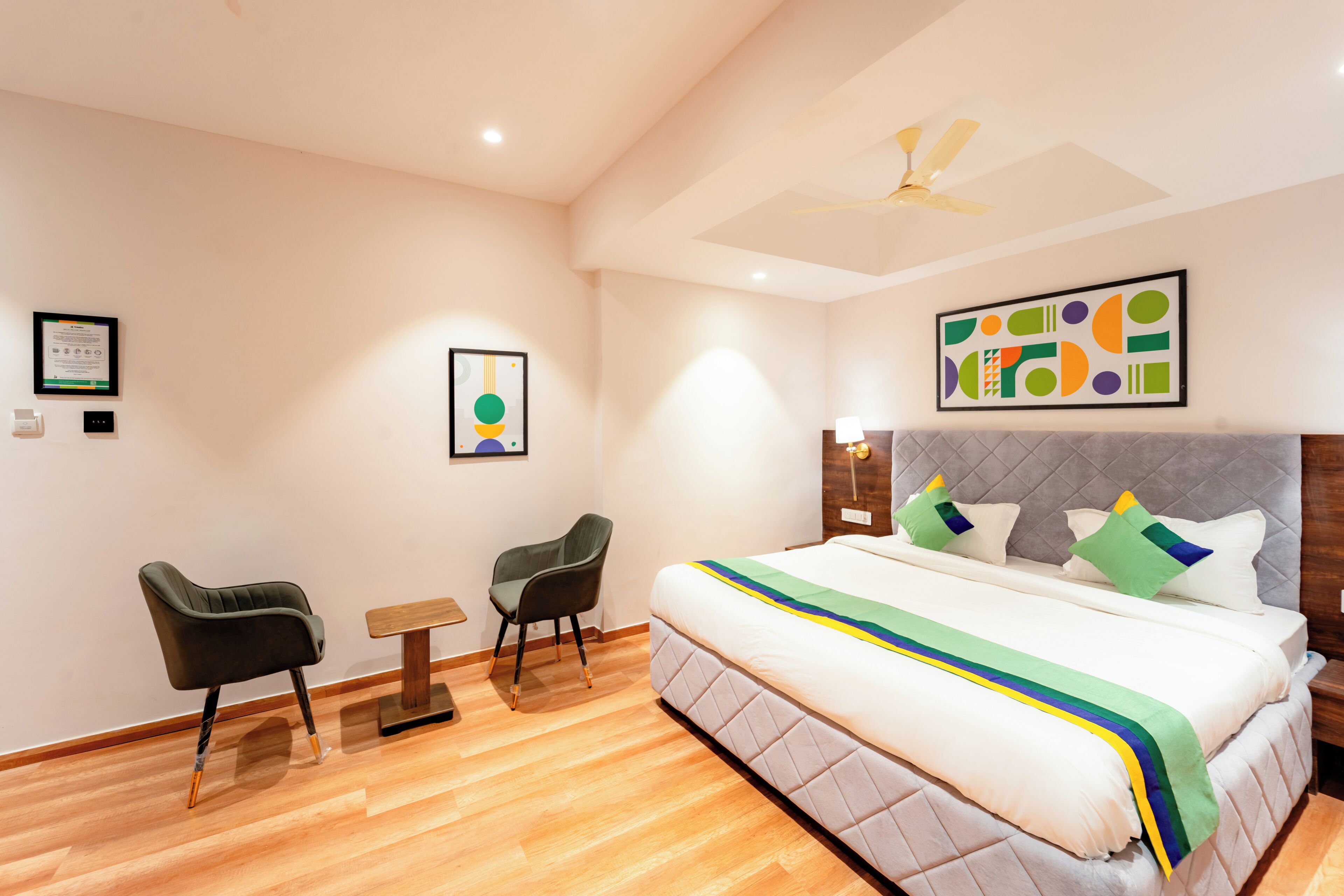 A modern hotel room with a large bed, two chairs, a small table, and artwork on the walls.