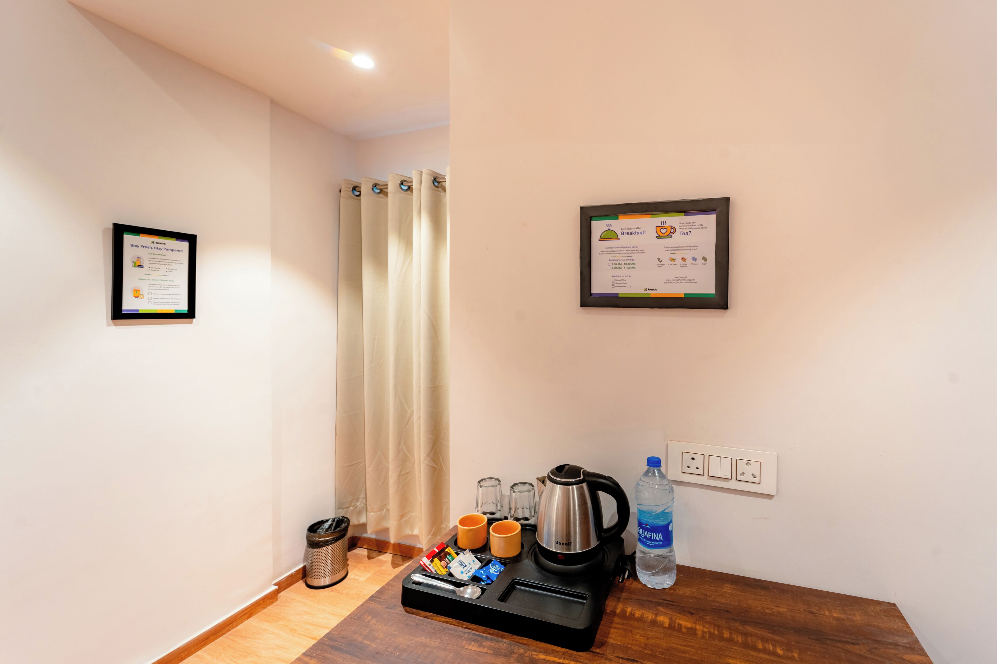 Une chambre d’hôtel avec une table en bois, une bouilloire, des tasses, une bouteille d’eau et une poubelle. Deux certificats encadrés sont accrochés au mur.