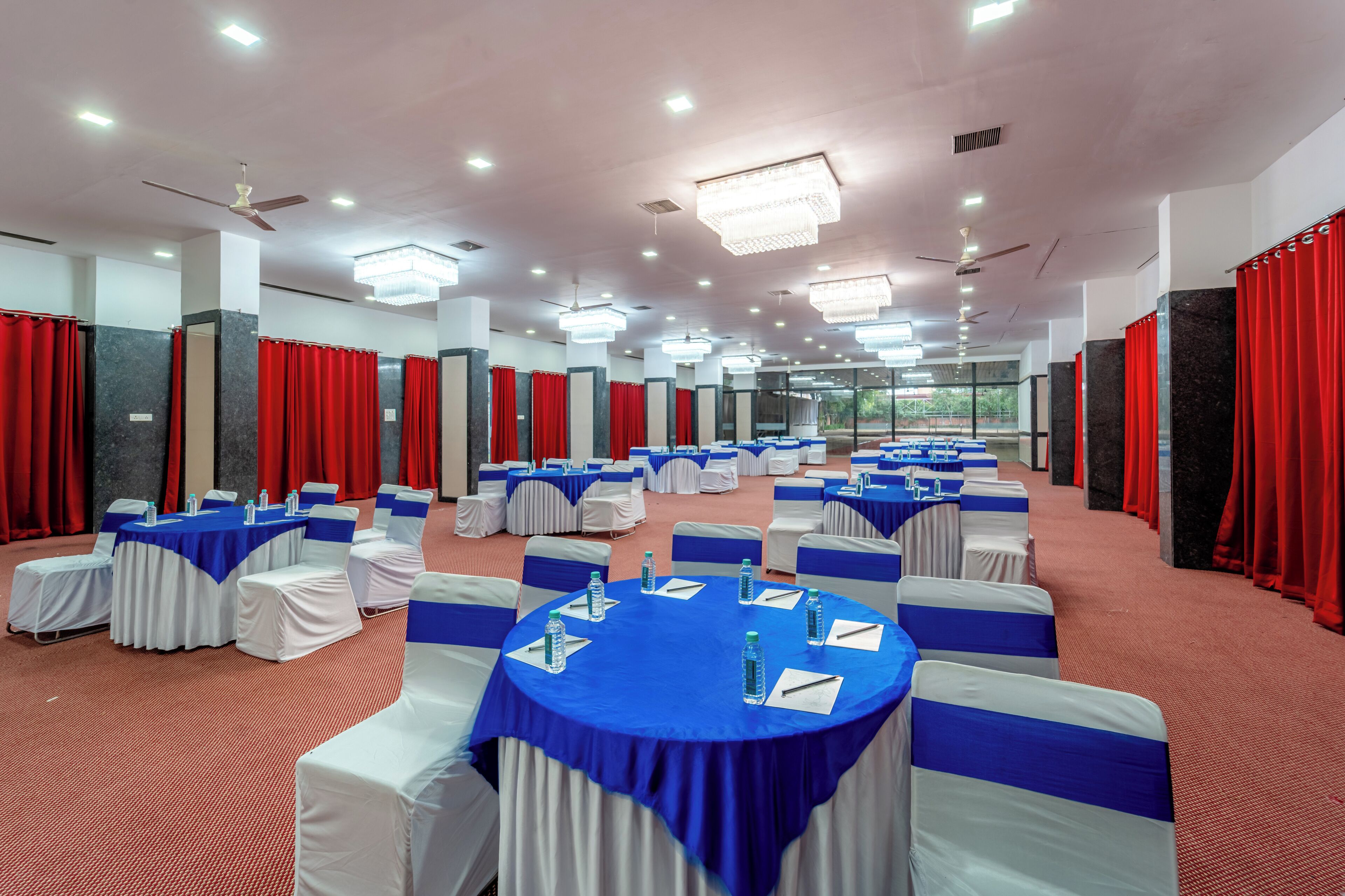 Un'ampia sala conferenze con tavoli rotondi, sedie bianche decorate con nastri blu, tende rosse e lampadari.