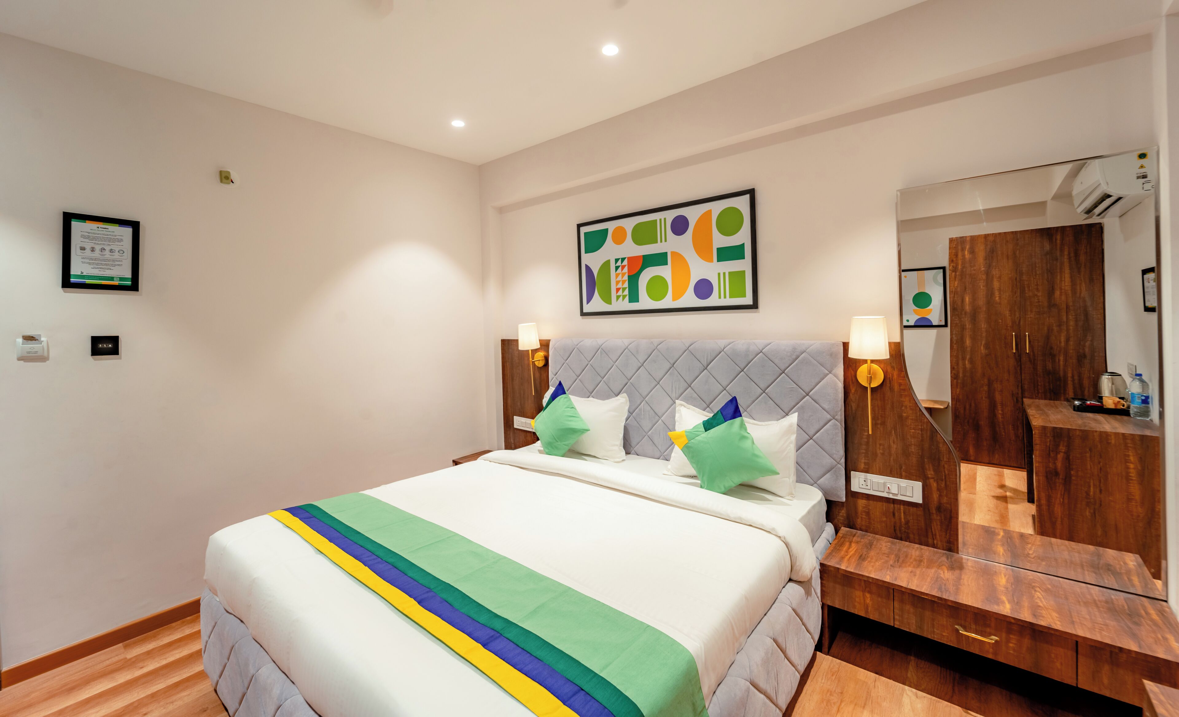 A hotel room with a large bed, a wooden nightstand, a mirror, and a wall-mounted artwork.
