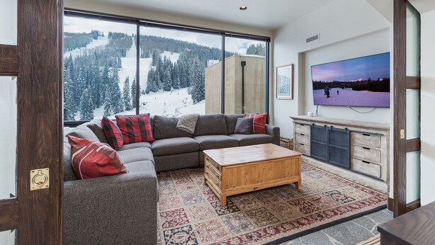 A living room with a large window, a sofa, a coffee table, a TV, and a cabinet. - Ski in/Out 7th Floor Penthouse at Purg - Huge Deck w/ Mountain/Slope Views/AC (Durango)