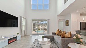 Ein geräumiges Wohnzimmer mit grauem Sofa, einem Flachbildfernseher, einem Kamin und Blick auf einen Swimmingpool.