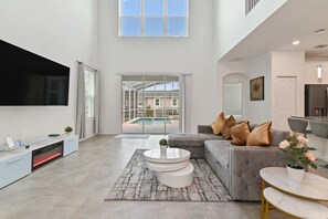 A spacious living room with a grey sofa, a flat-screen TV, a fireplace, and a view of a swimming pool.