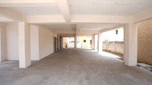 A long, narrow corridor with white walls and a concrete floor, leading to an open area with a view of a building in the distance. - Aishwaryam Service Apartment (Chennai)