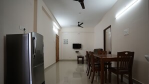 A modern kitchen with a stainless steel refrigerator, a dining table with chairs, a ceiling fan, and a flat-screen TV mounted on the wall. - Aishwaryam Service Apartment (Chennai)