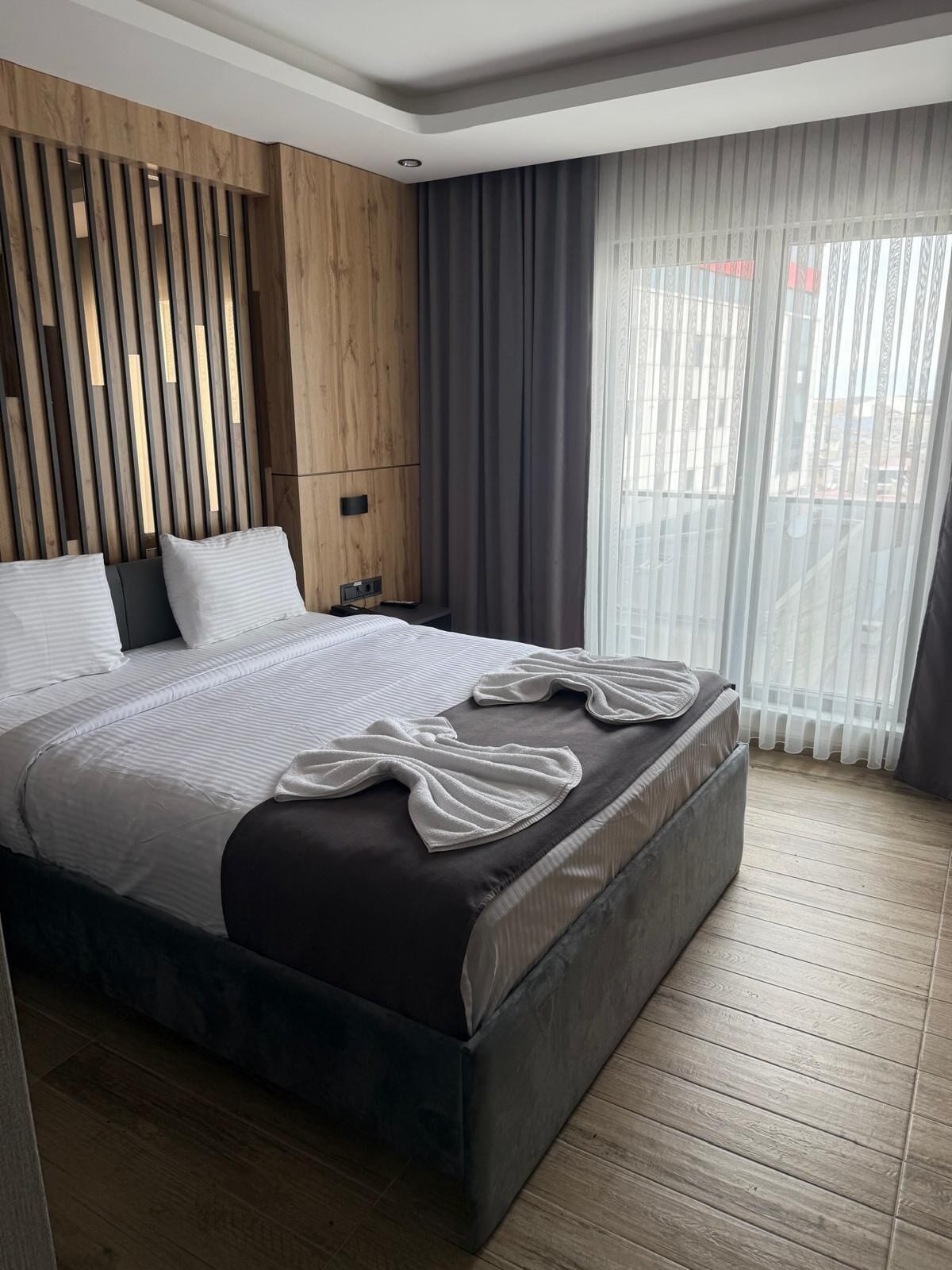 A modern bedroom with a large bed, wooden panel wall, and a view of the city through sheer curtains.