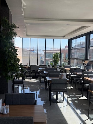 Un espace de détente intérieur moderne, doté de tables et de chaises, de grandes fenêtres et offrant une vue sur les bâtiments et les montagnes.