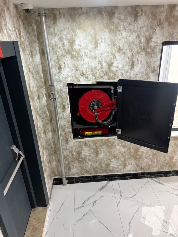 A room with a marble floor, a red fire hose reel mounted on the wall, and a black door. - WORLD SAY HOTEL (Van)