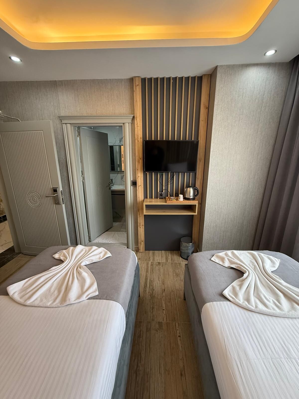 A hotel room with two beds, a TV mounted on a wooden panel, a small shelf with a kettle, and a door leading to a bathroom.
