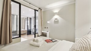 Una habitación de hotel moderna con una cama grande, una mesita de noche, una lámpara y vistas de la habitación contigua a través de una puerta corredera.