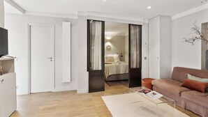 Una sala moderna con un sofá, una mesa de centro y un televisor. Hay una puerta que da a un dormitorio con una cama y una mesita de noche.