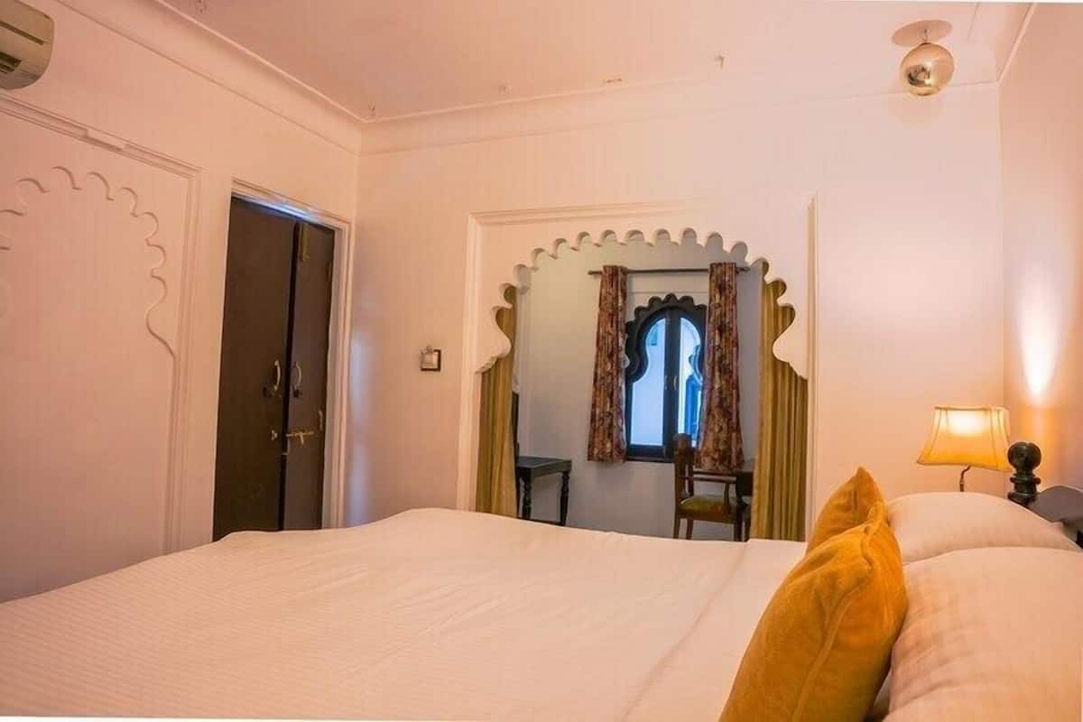 A hotel room with a bed, a window with curtains, a chair, a table, and a lamp.
