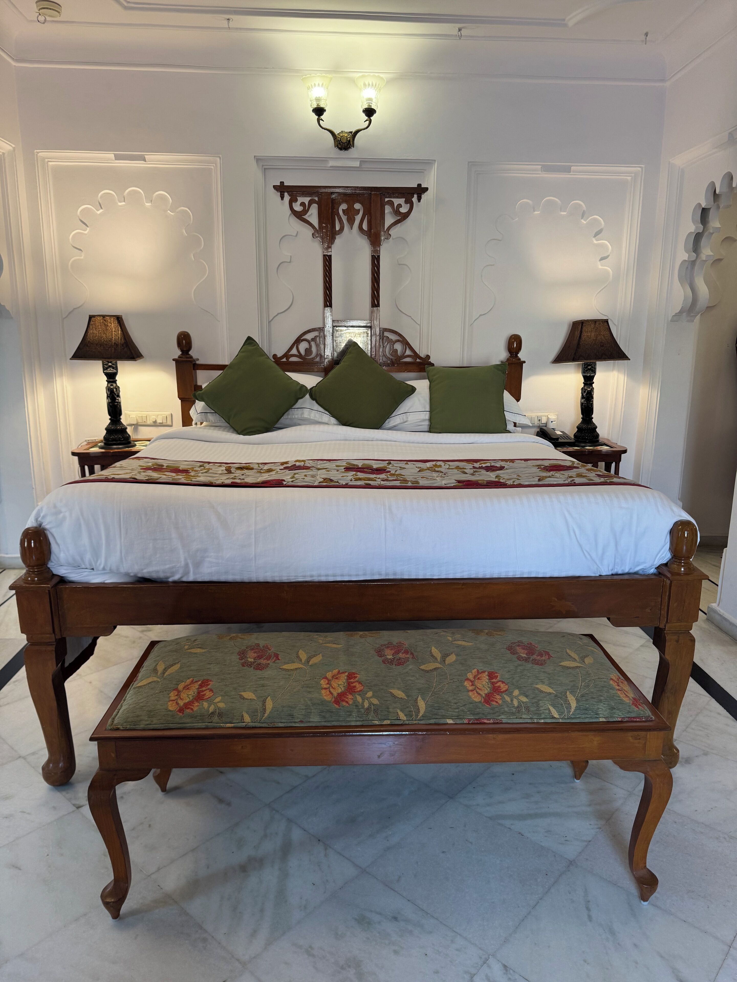 A classic four-poster bed with a floral bedspread and green pillows, flanked by two bedside tables with lamps, in a room with white walls and a decorative ceiling light fixture.
