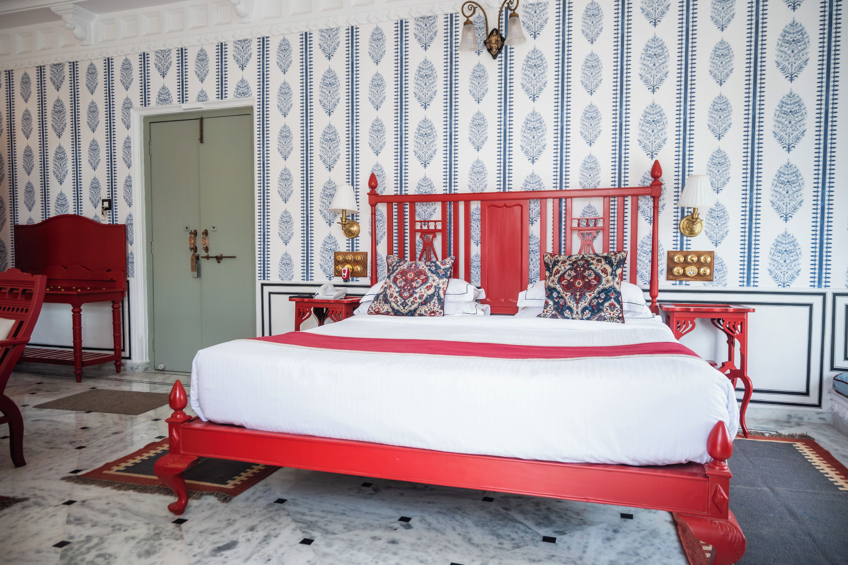 A room with a red bed, patterned wallpaper, a door, and a red chair.