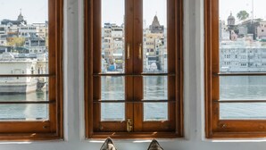 A room with three wooden-framed windows with colorful stained glass, a bench with cushions, and a view of a cityscape and water. - Hotel Amet Haveli (Udaipur)