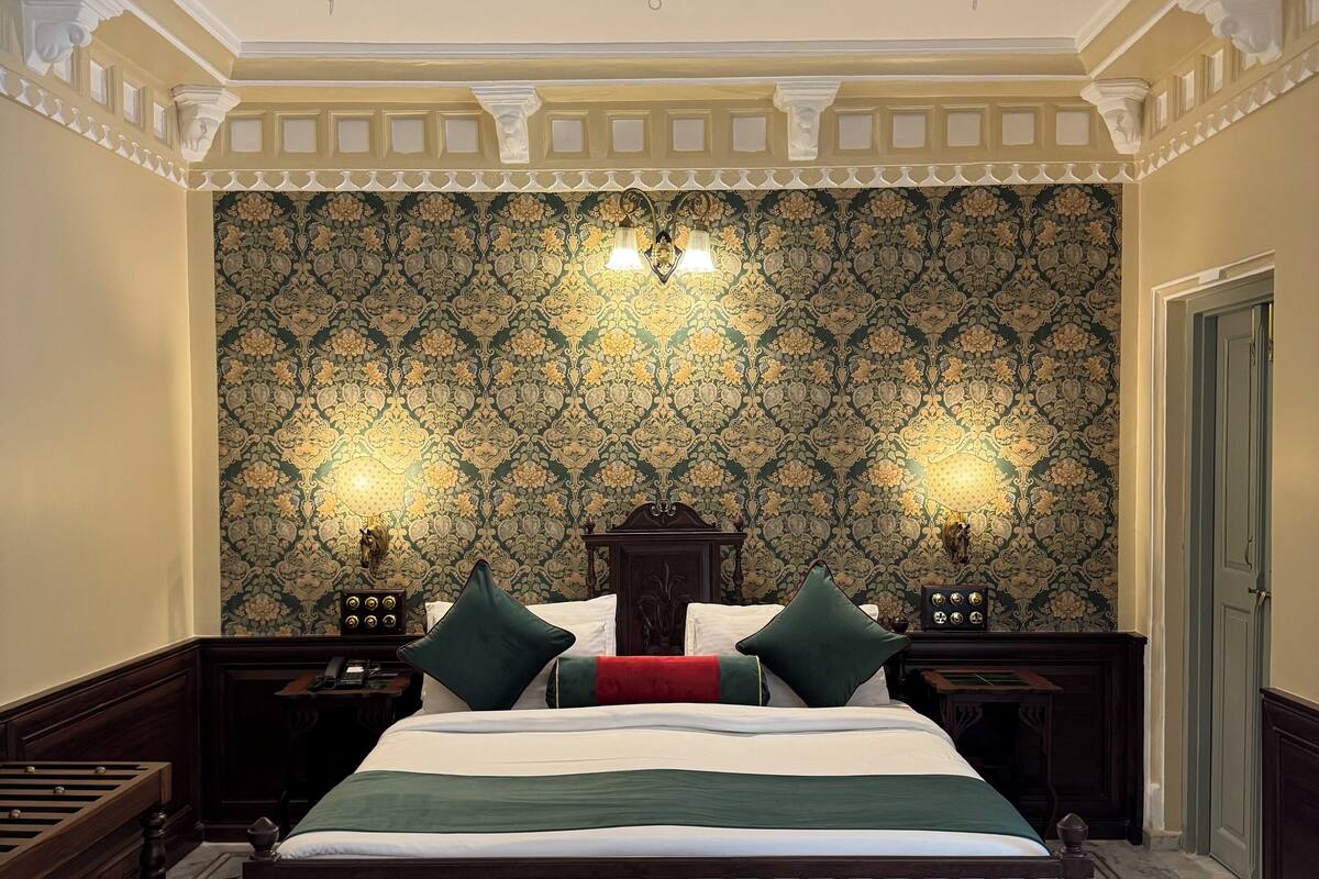 A bedroom with a large bed, a ceiling fan, ornate wallpaper, and a wooden nightstand.