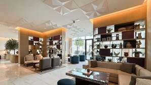 A modern living room with a sofa, armchairs, and a coffee table. There are shelves with decorative items and a large window providing a view of the city.
