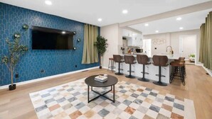 A modern living room with a large TV, a coffee table, and a patterned rug. There are bar stools and a kitchen area visible in the background. - Walk to OSU: Relaxing Retreat with Hot Tub (Columbus)