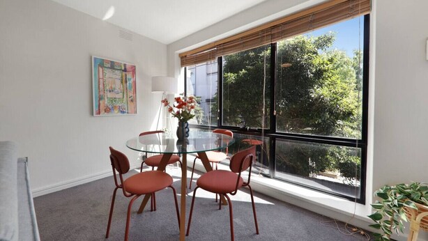 Un coin repas avec une table en verre, des chaises rouges, un vase rempli de fleurs et une grande fenêtre donnant sur des arbres.