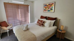 Une chambre bien rangée, avec un lit, une chaise, une table de chevet et une fenêtre équipée de stores.