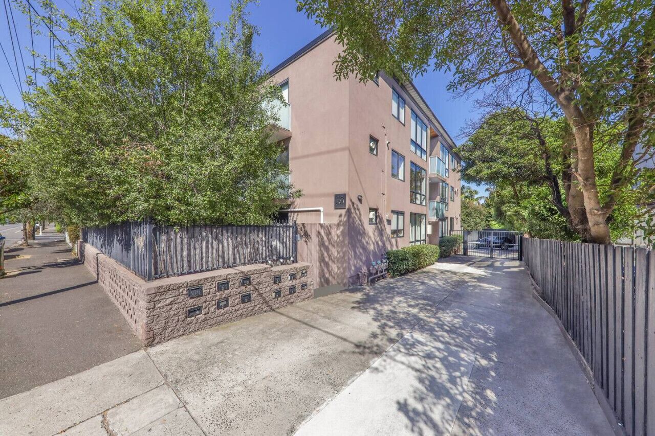 A multi-story residential building with a stone facade, a gated entrance, and a sidewalk leading to a parking area.