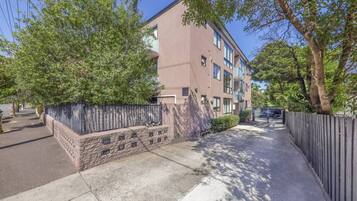 A multi-story residential building with a stone facade, a gated entrance, and a sidewalk leading to a parking area.