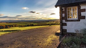 A traditional stone house with a gravel driveway and a view of rolling hills and fields.