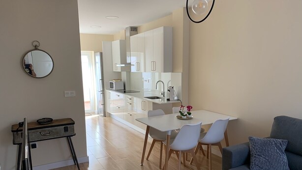 A modern living room with a grey sofa, a dining table with white chairs, and a kitchen area with white cabinets and appliances.