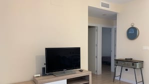 A modern living room with a flat-screen TV on a wooden stand, a small table with a bowl, and a wardrobe in the background.