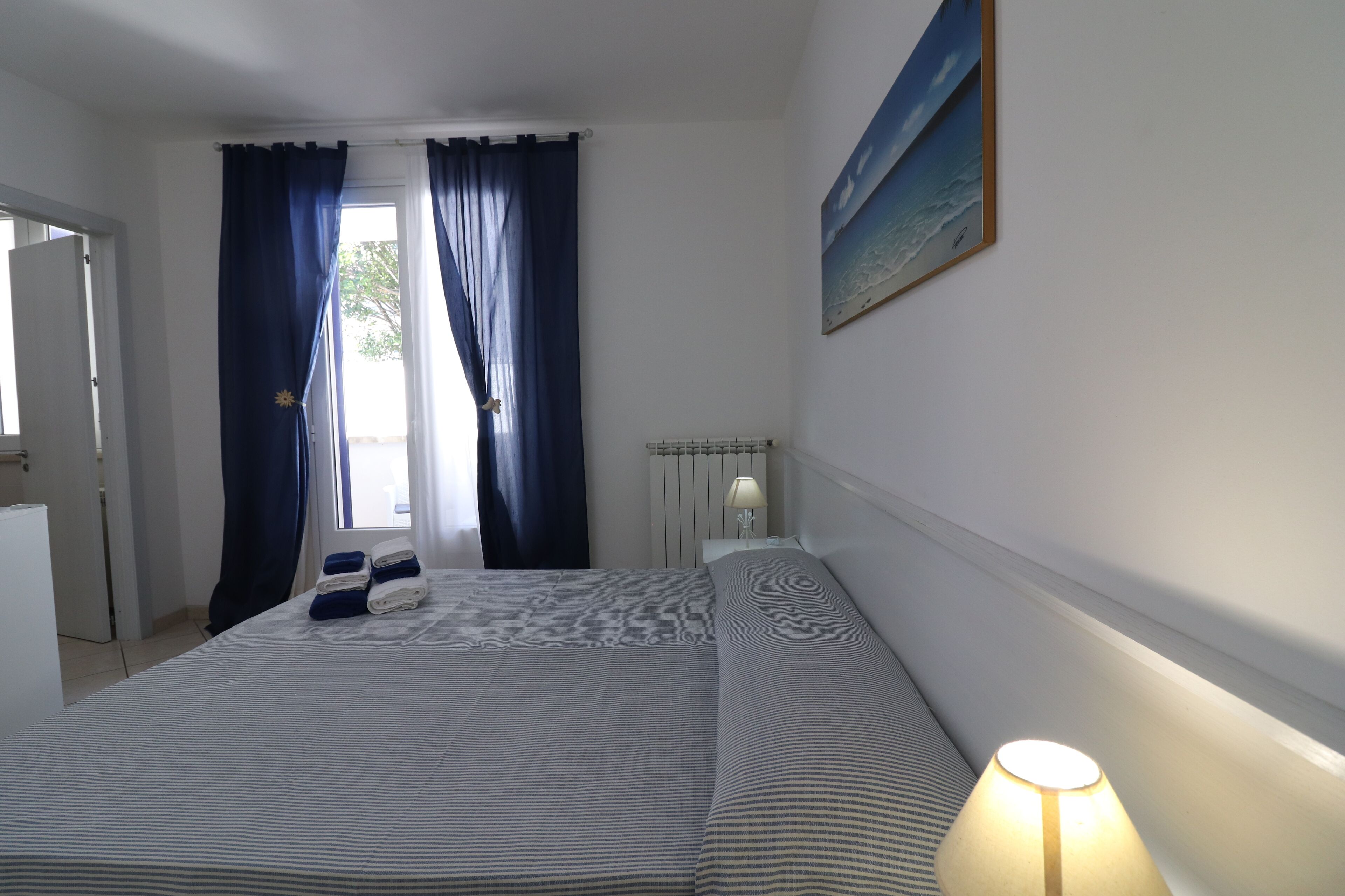 A bedroom with a bed, curtains, a lamp, a painting, and a radiator.