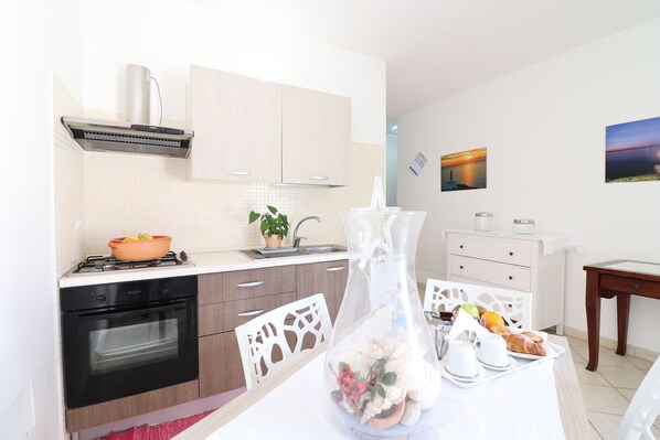 A modern kitchen with a gas stove, oven, and white cabinets. There is a dining table with chairs, a vase with seashells, and a painting on the wall.
