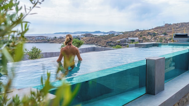 A modern infinity pool with clear glass edges, overlooking a coastal landscape with buildings and hills. - 2 Bedroom Villa with Private  Glass Pool & Shared Infinity Pool, Gym, Pool Bar (Mykonos)