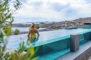 A modern infinity pool with clear glass edges, overlooking a coastal landscape with buildings and hills.