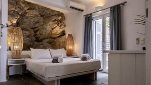 A bedroom with a stone wall, a bed with white bedding, two bedside lamps, and a window with curtains. - 2 Bedroom Villa with Private  Glass Pool & Shared Infinity Pool, Gym, Pool Bar (Mykonos)