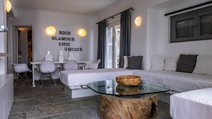 A modern hotel room with a stone floor, a glass coffee table, a sofa, a bed, and a desk with chairs. - 2 Bedroom Villa with Private  Glass Pool & Shared Infinity Pool, Gym, Pool Bar (Mykonos)