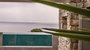 A modern building with a stone facade and a large glass window overlooking a body of water. - 2 Bedroom Villa with Private  Glass Pool & Shared Infinity Pool, Gym, Pool Bar (Mykonos)