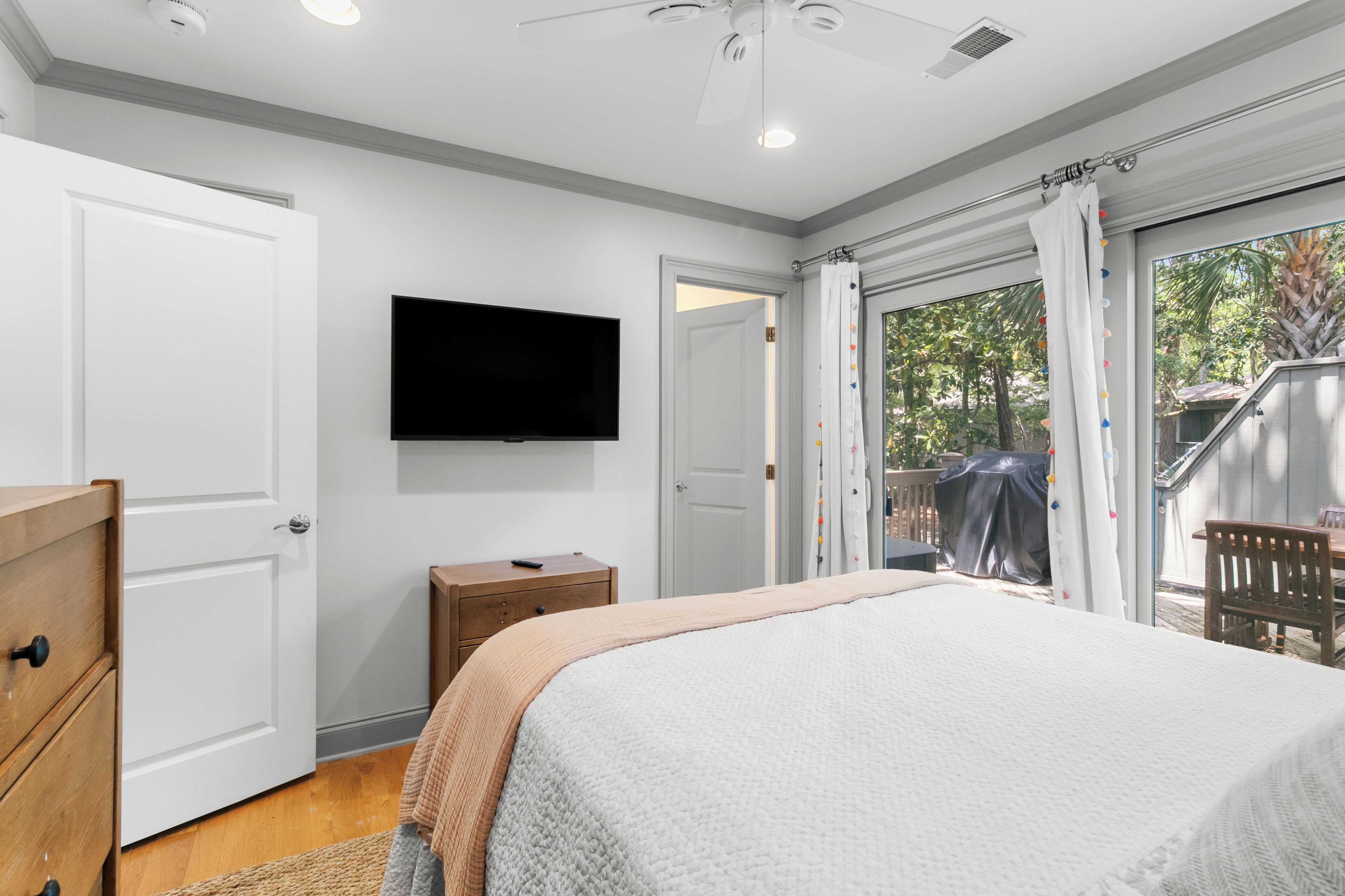 A bedroom with a bed, a TV mounted on the wall, a wooden nightstand, and a door leading to a balcony.