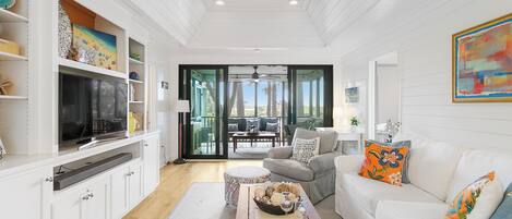 A spacious living room with a large TV, white furniture, and a view to the outdoors.