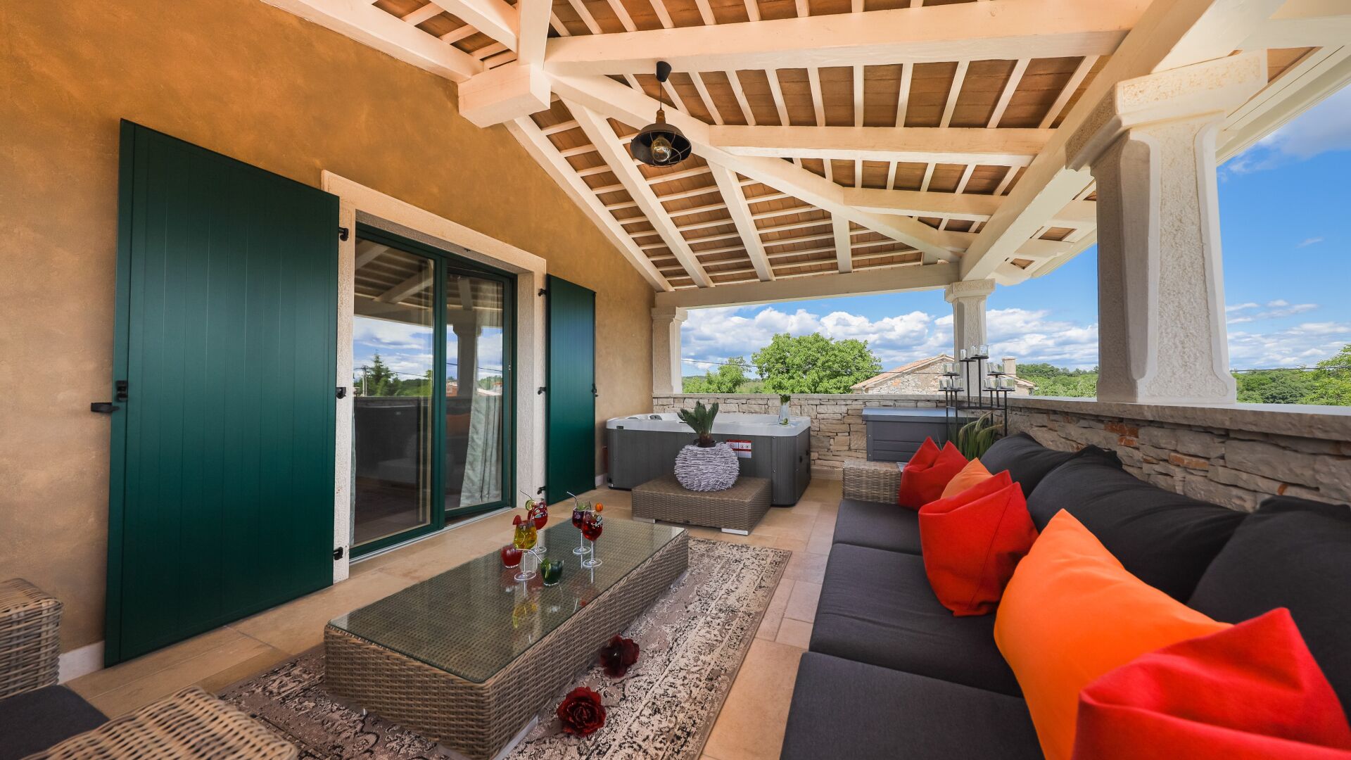 An outdoor patio with a glass door leading to a covered area, a hot tub, and a seating area with cushions.