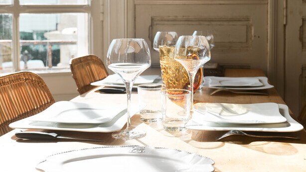 Una mesa de comedor dispuesta con platos, cubiertos y vasos, con una silla de mimbre a la vista.