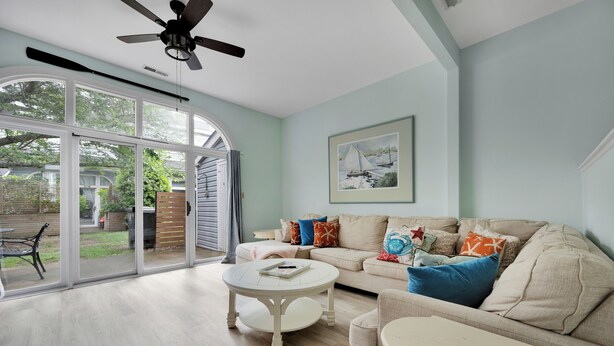A spacious living room with a large sliding glass door leading to a backyard, a ceiling fan, a beige sofa with colorful cushions, and a white coffee table. - In-town five bedroom townhouse - close to the beach! (Lewes)