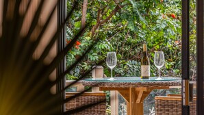 A table with a bottle of wine and two glasses, set against a backdrop of lush greenery.