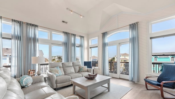 A spacious living room with a large sofa, a coffee table, and a view of the outside. - Blue Bomber: Oceanside | Pool/Hot Tub | Dogs OK (Corolla)