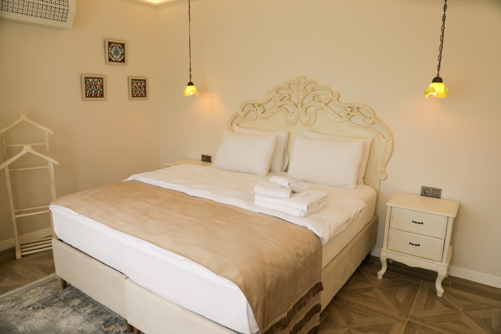 A neatly made bed with a beige bedspread, white pillows, and folded towels on a wooden floor. There is a small bedside table with a lamp and a clock.