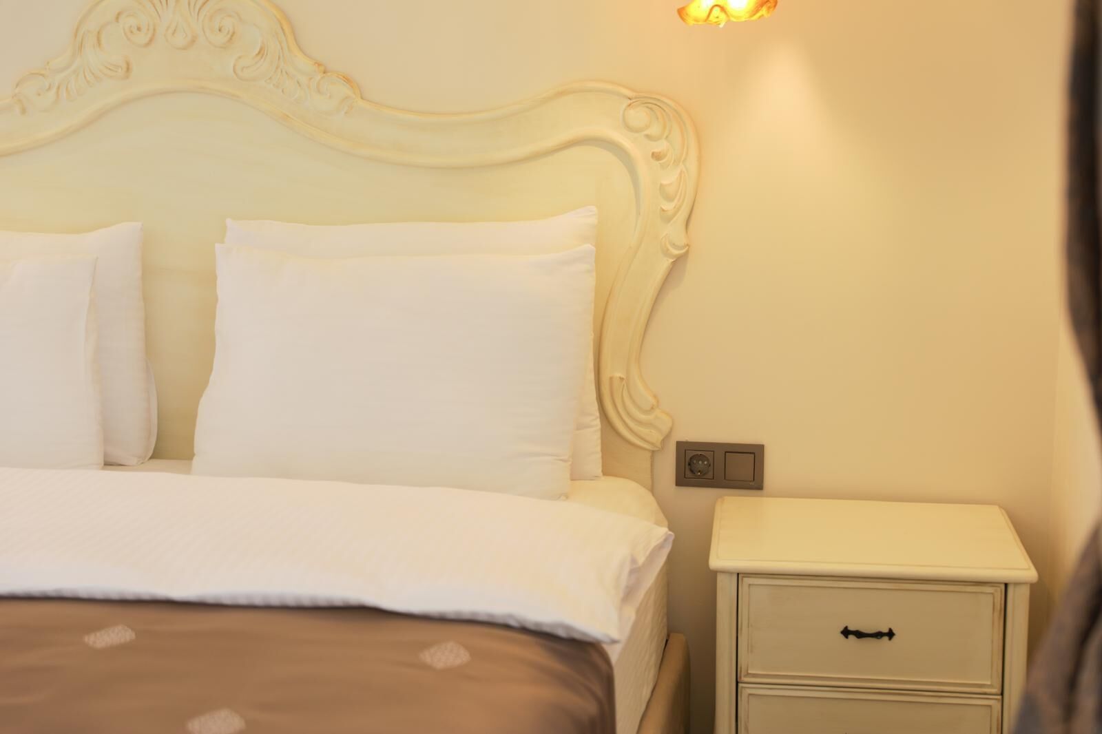 A neatly made bed with white linens, a cream-colored headboard, and a bedside table with a drawer.