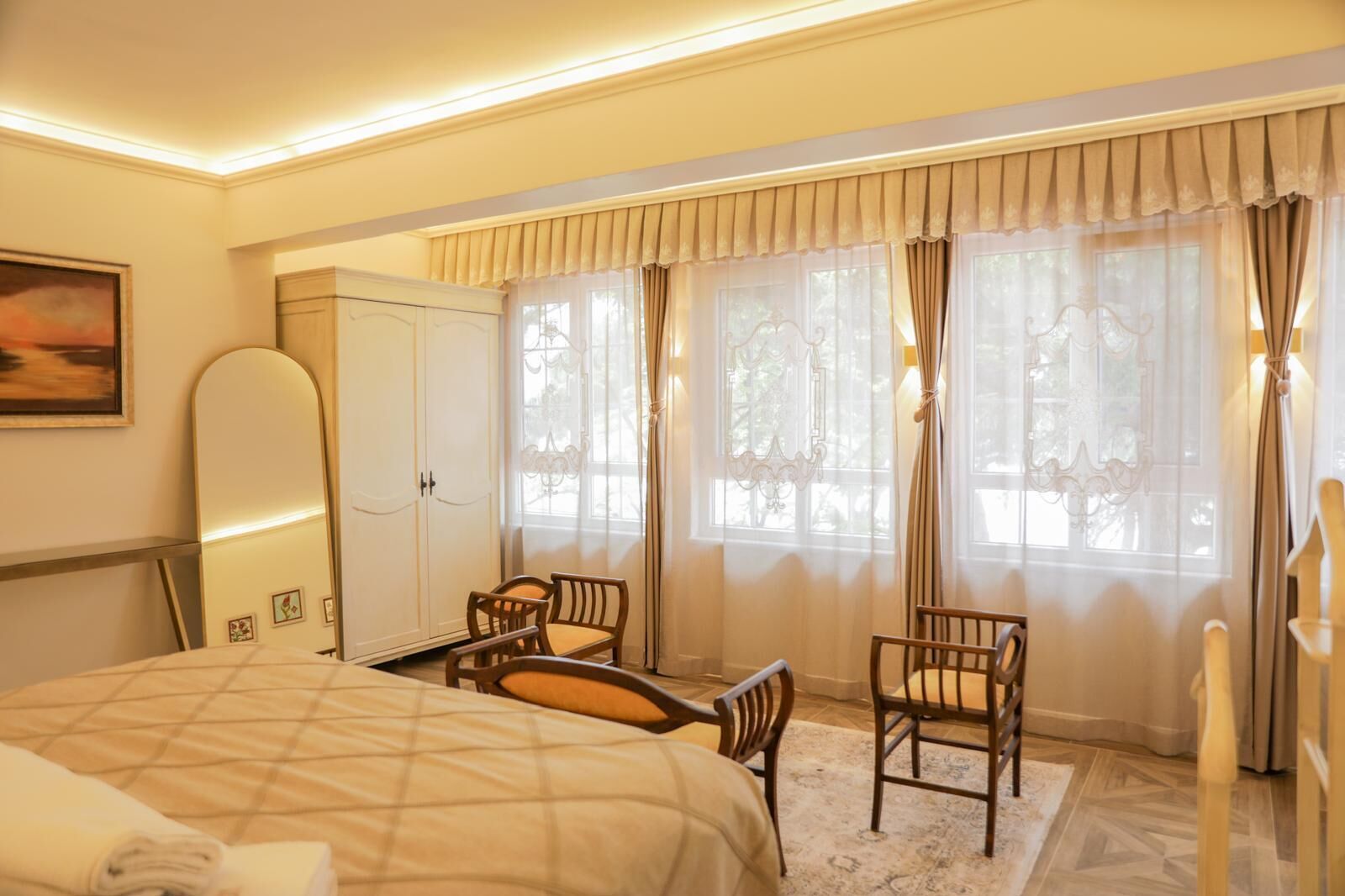 Une chambre d’hôtel comprenant un lit, un bureau, deux chaises, une armoire et de grandes fenêtres avec des rideaux.
